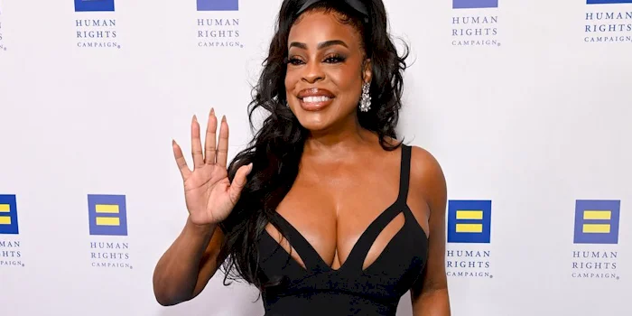 Close up of Niecy Nash in a fitted black gown with sheer accents, smiling and waving in front of a backdrop with repeated Human Rights Campaign logos.