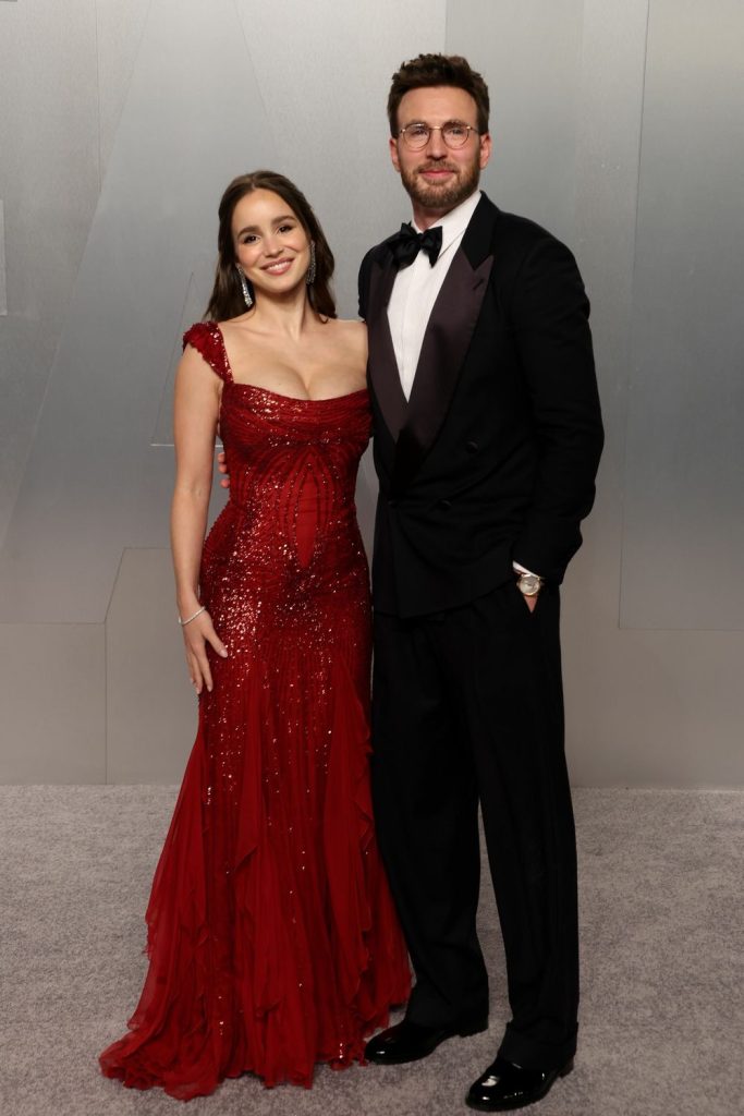 A man and a woman smile for the cameras. The woman is in a long red gown and the man is wearing a black tux.