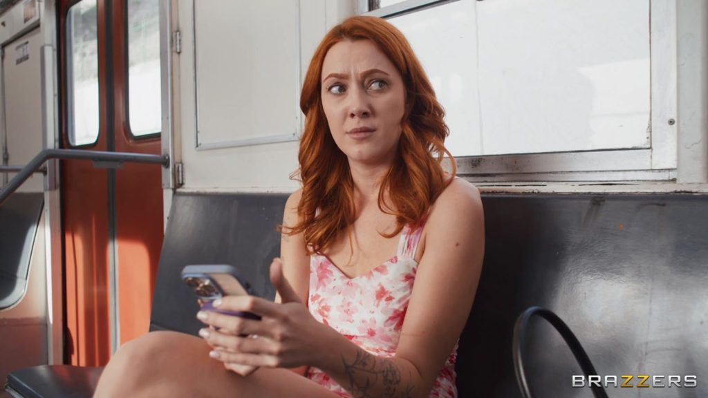 A redhead woman sitting on a train is looking to her side. She looks somewhat disturbed.