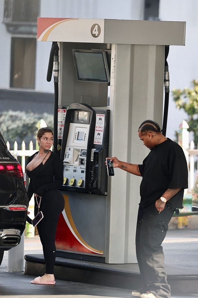 A woman in black leggings and a top stands by an SUV at a gas pump while a man in black clothes holds a can, possibly spraying or showing it, near the pump. Both appear to be in conversation at the gas station.