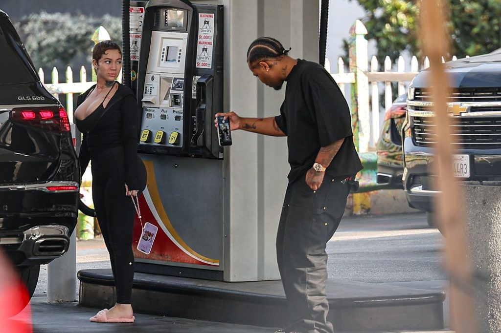 A woman in black clothes and pink slippers stands by a gas pump while a man in black clothes takes a photo of her with his phone at a gas station. Two black vehicles are parked nearby.