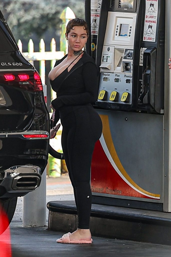 A woman in black leggings and a long-sleeve top pumps gas into a black SUV at a gas station. She is wearing pink slippers and looking toward the camera, with gas pumps and warning signs visible in the background.