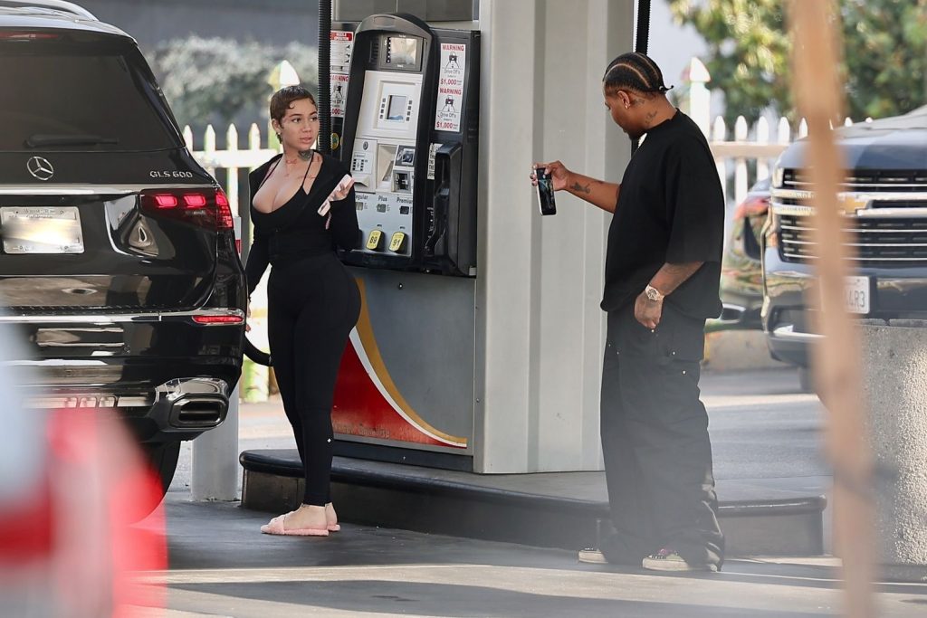 A woman in black clothing pumps gas barefoot at a station while a man in black holds up a phone and appears to take her photo or video. A black SUV is next to them at the gas pump.