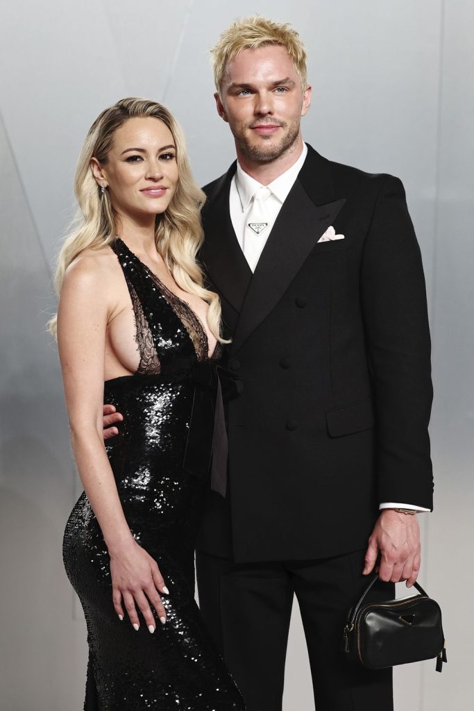 A man and a woman stand close together at a red carpet event. The woman wears a sleek black dress and the man wears a suit.