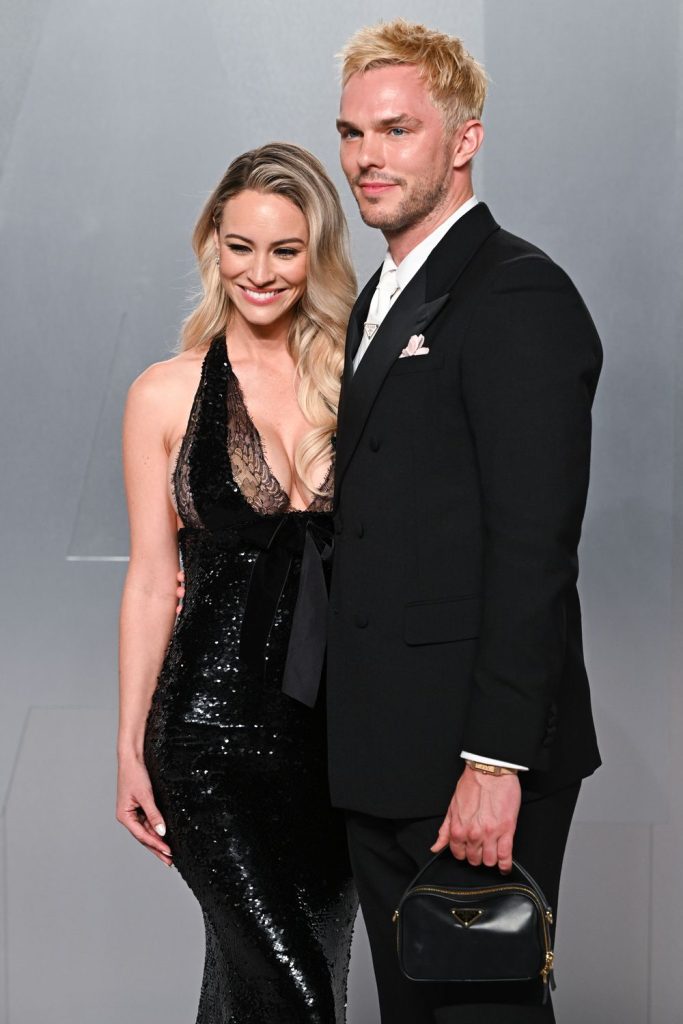A man and a woman stand close together at a red carpet event. The woman smiles and is turned facing the camera and the man is at a side angle.