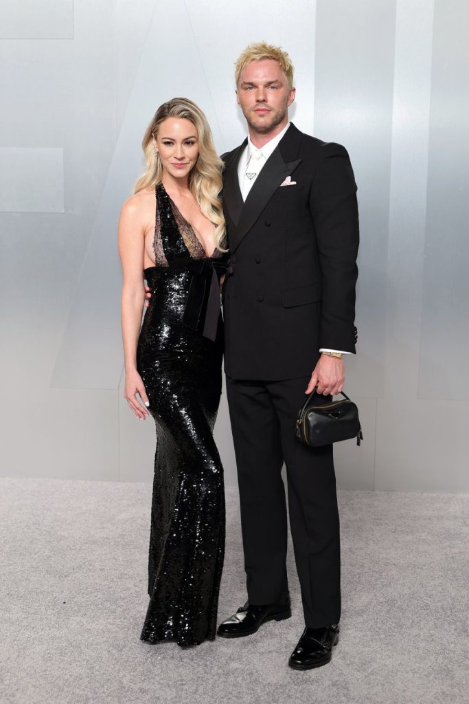 Far away view of a man and a woman stand close together at a red carpet event. The woman wears a sleek black dress and the man wears a suit.