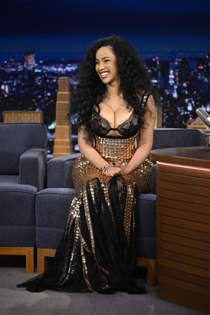 Cardi B with long curly hair wearing a glamorous, sequined black and gold dress sits smiling on a talk show set with a cityscape backdrop.