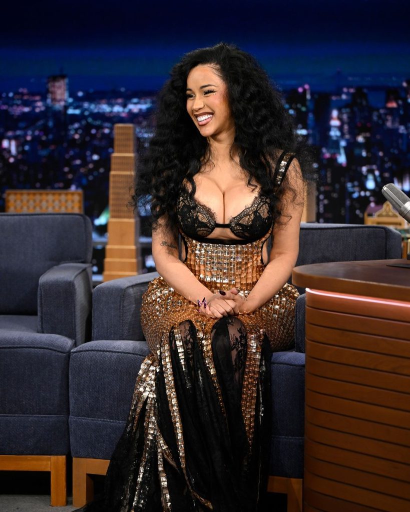 Cardi B with long curly hair in a glamorous black and gold dress sits smiling on a talk show set, with cityscape lights in the background and a blue sofa and a desk nearby.