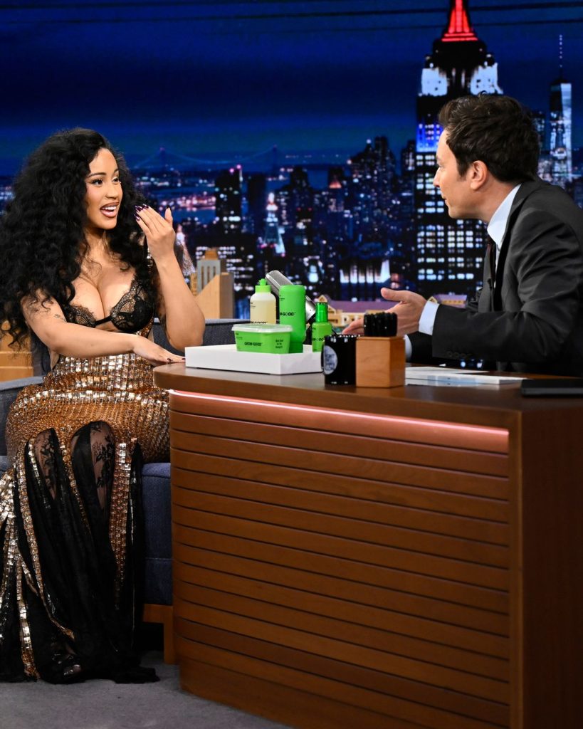 Cardi B in a sparkling gold and black dress talks animatedly to a man in a suit across a desk on a late-night talk show set, with a city skyline backdrop behind them.