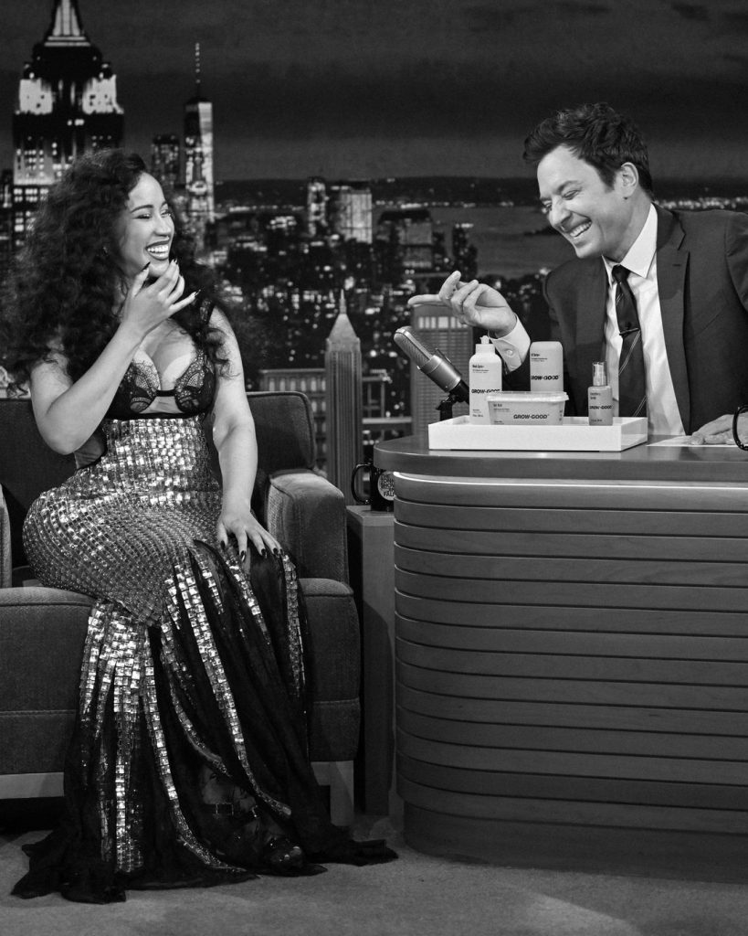 A woman in a sparkly dress laughs while sitting on a talk show set, facing a male host in a suit who is smiling as he talks and gestures, with a city skyline backdrop behind them.