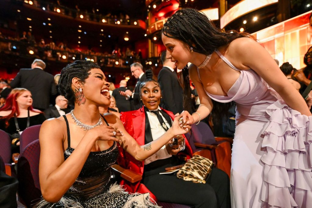A girl wearing lavender dress bending down to greet another woman sitting among an audience. The woman looks very happy. A lot of other people are in the background.