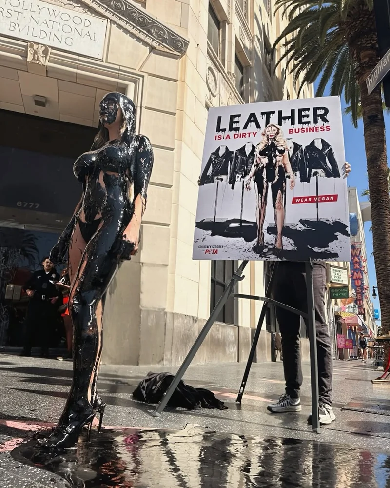 A woman standing on a public sidewalk wearing a bikini and drenched from head to toe with a thick black substance.