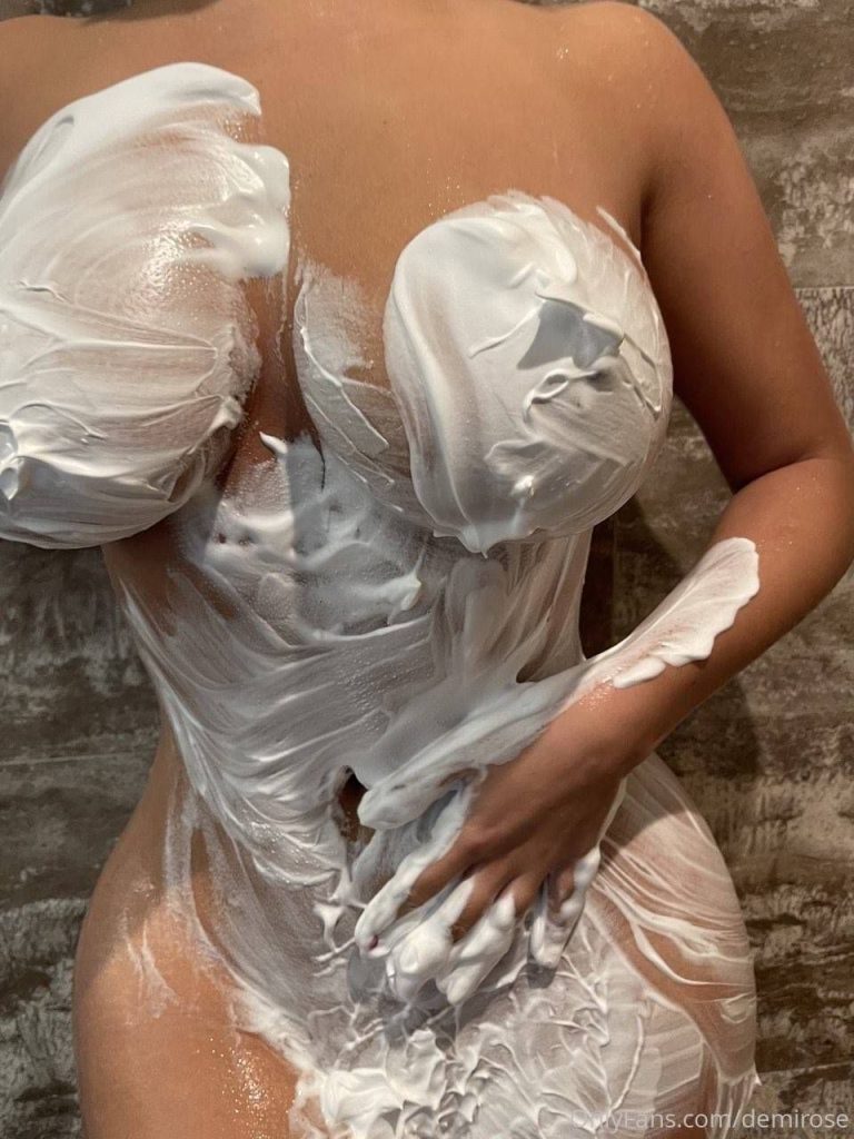 A person with a curvy figure standing against a tiled wall, covered in thick white foam or shaving cream over their chest, torso, and hand, with their head out of frame.