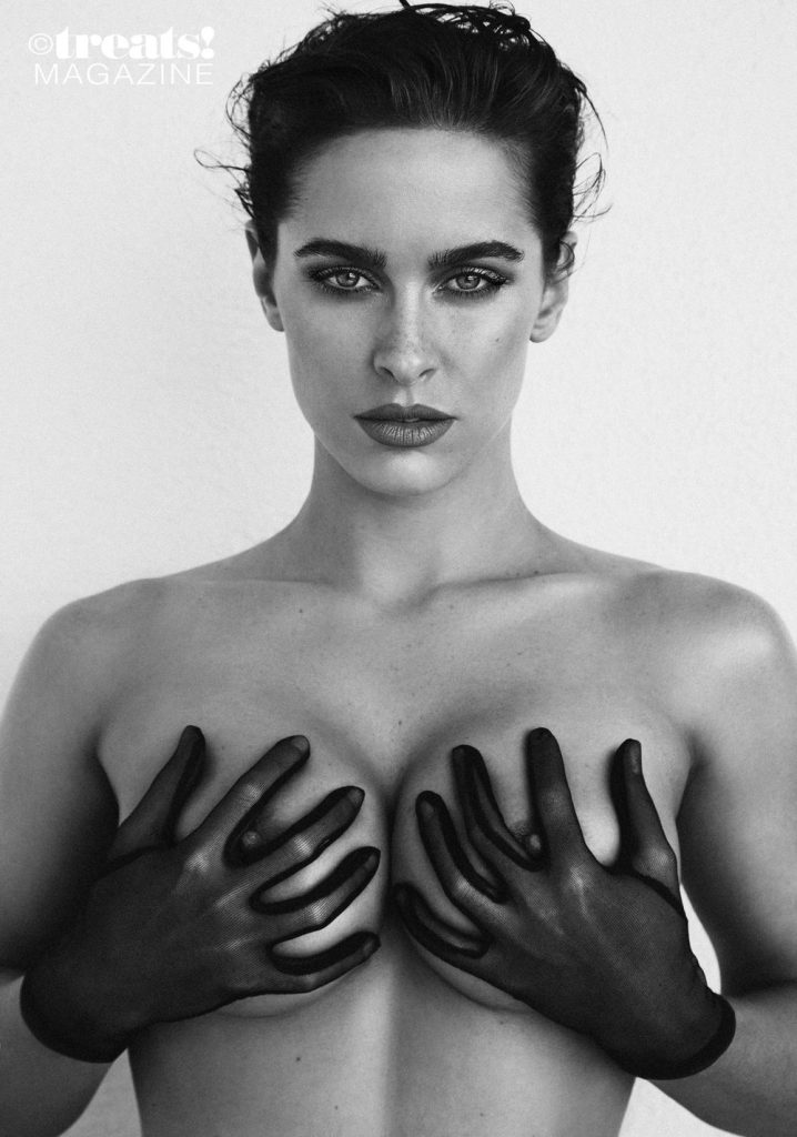 Black and white of a topless model wearing gloves and grabbing her breasts in front of a white background.