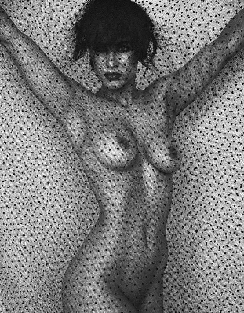 Black and white of a nude model wearing a hat and holding up a dotted sheer fabric in front of herself