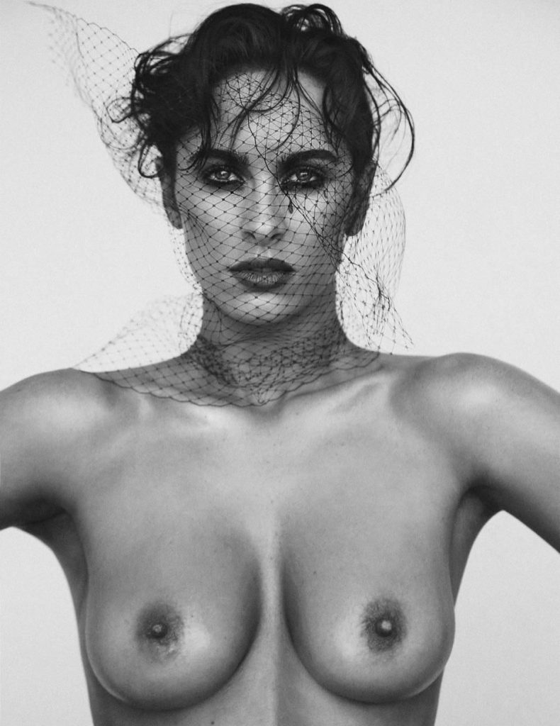 Black and white of a topless model with messy hair and makeup looking into the camera in front of a white background.