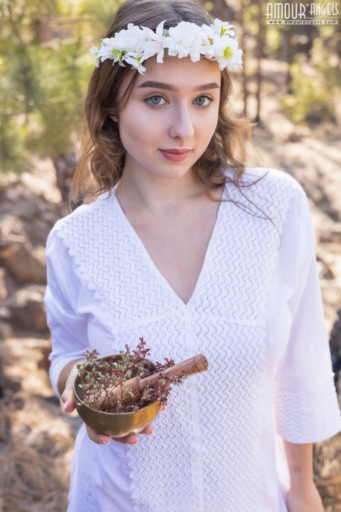 Close up of girl wearing flower crown and white dress in a forest. She is holding a small bowl and wood morsel in her hand.