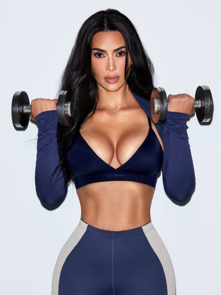 A woman with long dark hair, wearing a navy blue sports bra and matching leggings, holds a dumbbell in each hand at shoulder height, standing against a plain light background.
