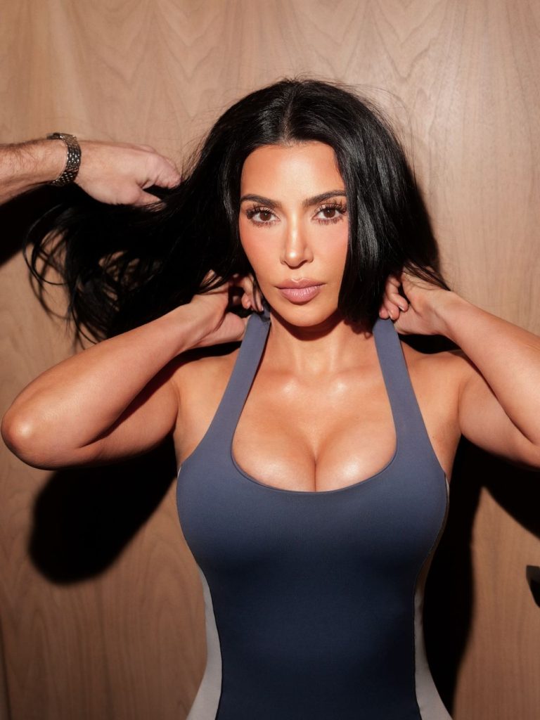 A woman with long dark hair and a fitted blue-gray sleeveless top poses in front of a wooden background while someone adjusts her hair from the left.