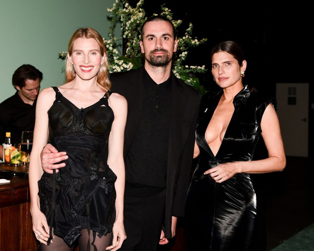 Three people pose together indoors in evening attire; two women in black dresses stand on either side of a man in a black suit. A floral arrangement is visible in the background, with a bar and bartender behind them.