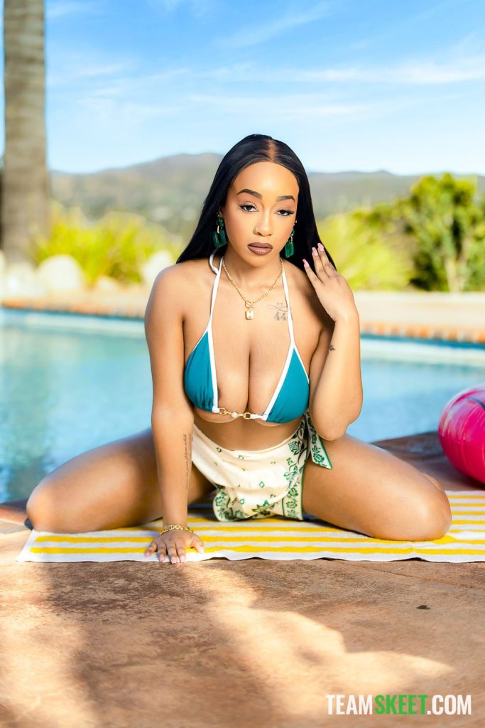 A black woman wearing a bikini and skirt is sitting on her knees and is leaning forward towards the camera. She is in front of a swimming pool and a green valley can be seen in the background.