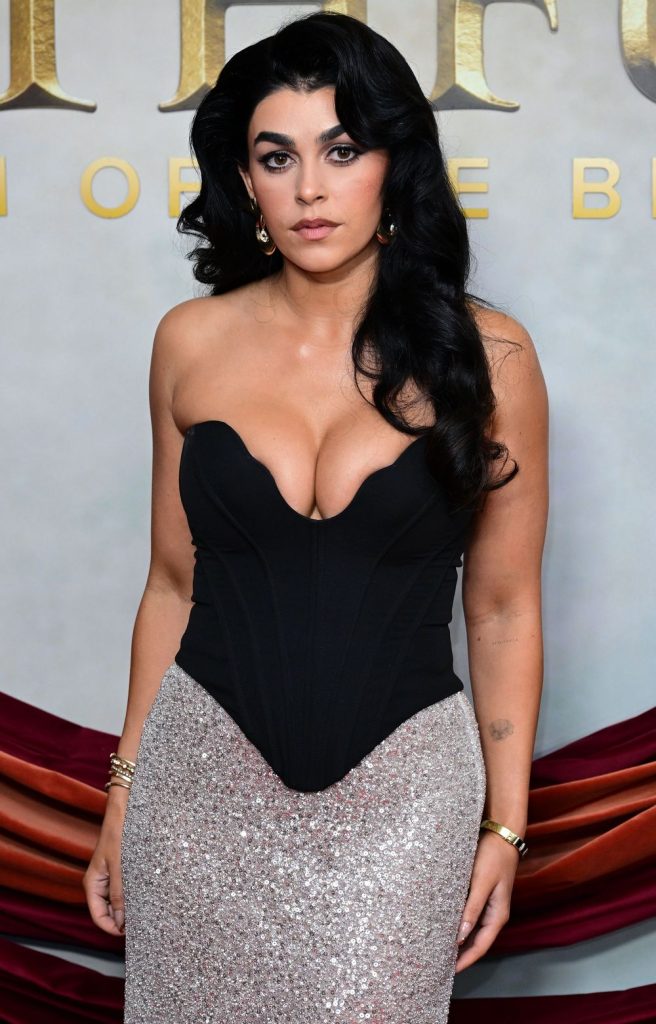 A woman with long, wavy black hair wears a strapless black and silver gown with a deep neckline. She stands in front of a light backdrop with gold lettering, looking directly at the camera with a neutral expression.