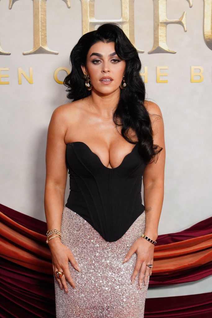 A woman with long, dark, wavy hair wears a strapless black and silver sparkling gown, standing in front of a backdrop with large gold letters and draped burgundy fabric.