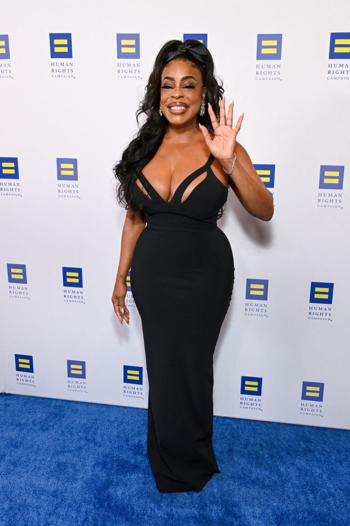 A black woman in a fitted black gown poses on a blue carpet, smiling and waving at the camera. The backdrop displays repeated logos for the Human Rights Campaign.