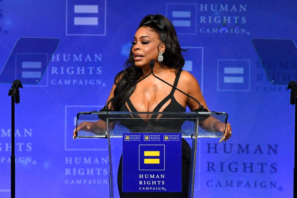 A woman with long dark hair speaks at a podium with the Human Rights Campaign logo, wearing a black dress, against a blue background with Human Rights Campaign repeated in white text.