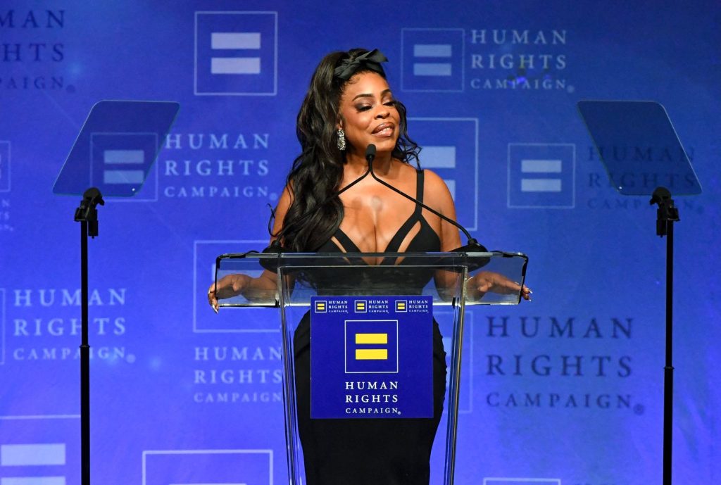 A black woman in a low cut dress speaks at a podium with the Human Rights Campaign logo, wearing a black dress, against a blue background with Human Rights Campaign repeated in white text.