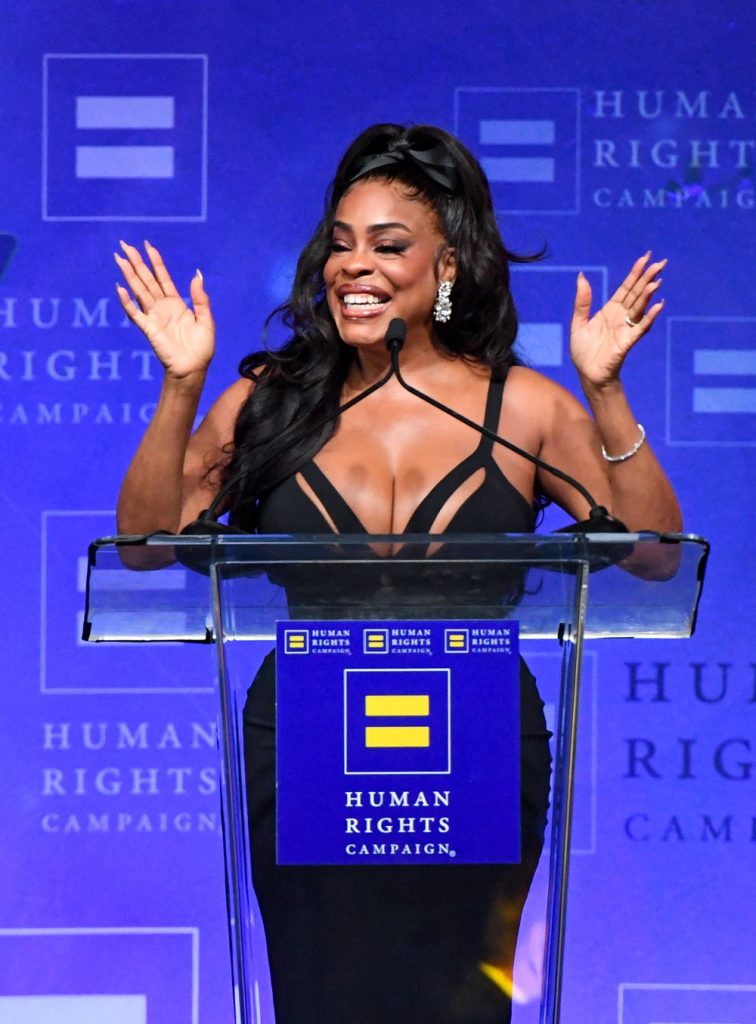 A black woman in a low cut dress throws her hands up at a podium with a blue background.
