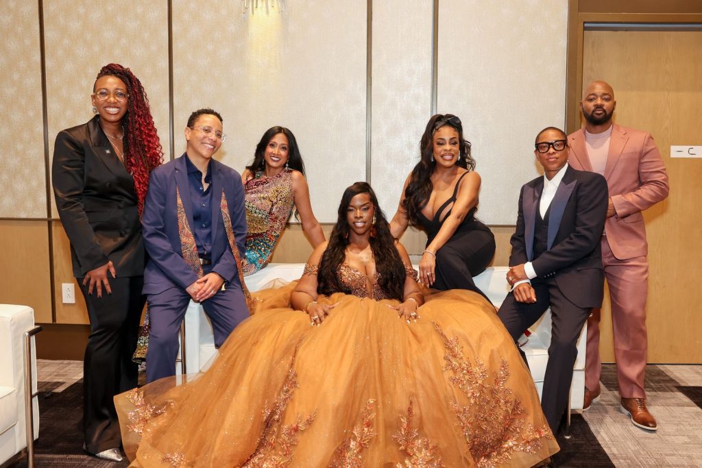 Seven people pose together indoors, with one person in the center seated in an elegant, flowing bronze gown. The others, dressed in suits and formal attire, stand or sit around her, smiling in a well-lit, modern room.