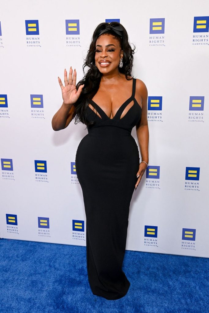 A woman in a fitted black gown with sheer accents stands on a blue carpet, smiling and waving in front of a backdrop with repeated Human Rights Campaign logos.