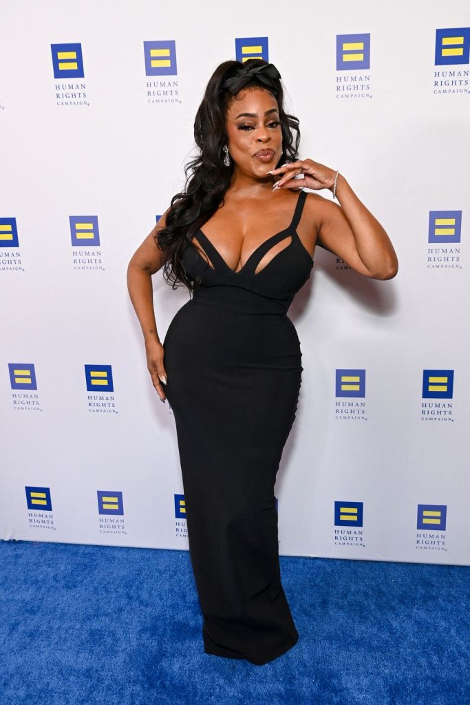 A black woman in a fitted black gown poses confidently on a blue carpet in front of a step-and-repeat backdrop with Human Rights Campaign logos. She has long wavy hair and accessorizes with earrings and rings.
