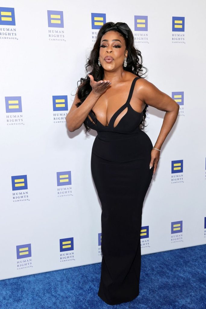 A woman in a form-fitting black dress blows a kiss while posing on a blue carpet in front of a backdrop with repeating Human Rights Campaign logos.