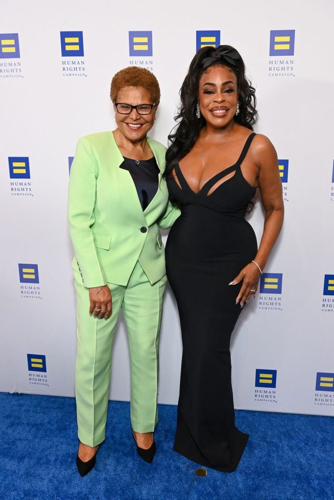 Two women stand smiling on a blue carpet in front of a Human Rights Campaign backdrop; one wears a light green suit and glasses, the other wears a fitted black gown with cutout details.