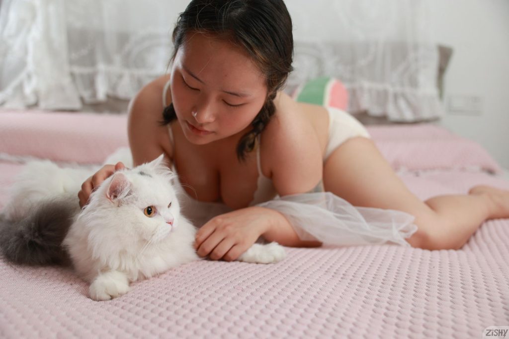 Asian woman in light-colored lingerie lies on a pink bed while gently petting a fluffy white cat with gray markings. The setting appears soft and cozy, with sheer curtains in the background.