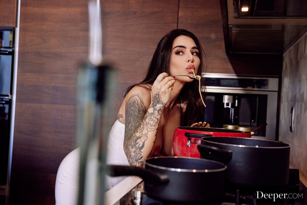 A woman with long dark hair and tattoos tastes spaghetti from a fork while cooking in a modern kitchen, standing near several pots on the stove.