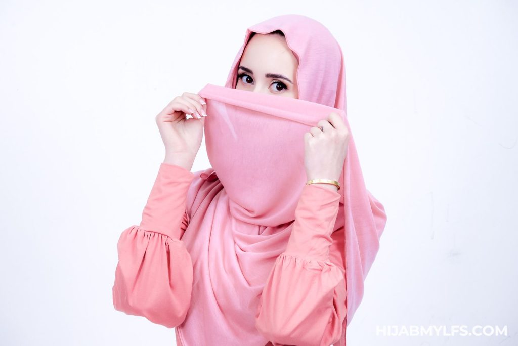A woman wearing a pink hijab and long-sleeved top covers the lower half of her face with her hijab, revealing only her eyes. The background is plain white.