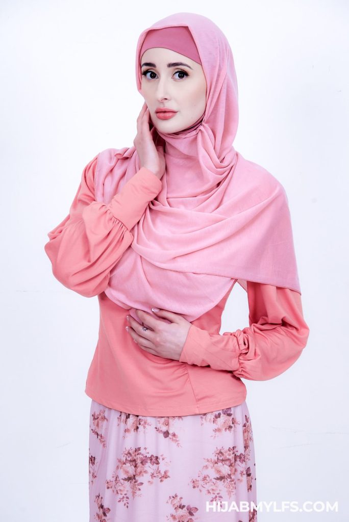 A woman wearing a light pink hijab, a long-sleeved peach blouse, and a floral pink skirt stands against a white background, gently touching her neck with one hand.