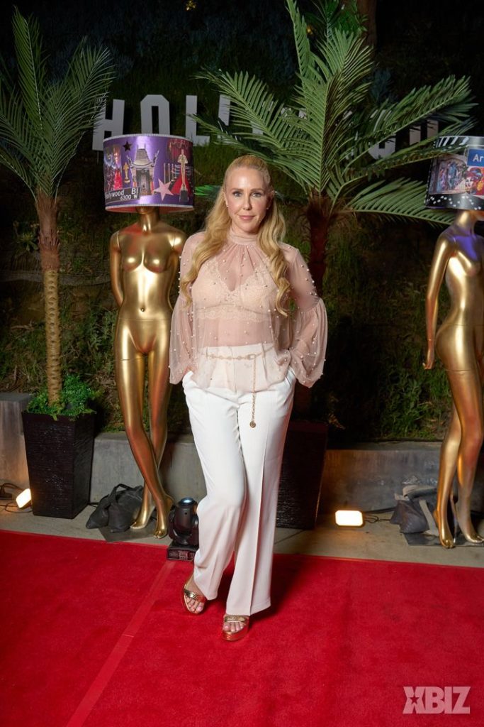 A woman with long blonde hair, wearing a sheer beige blouse and white pants, poses on a red carpet between two gold mannequins with lamp shades on their heads. Palm plants and the word HOLLYWOOD are in the background.