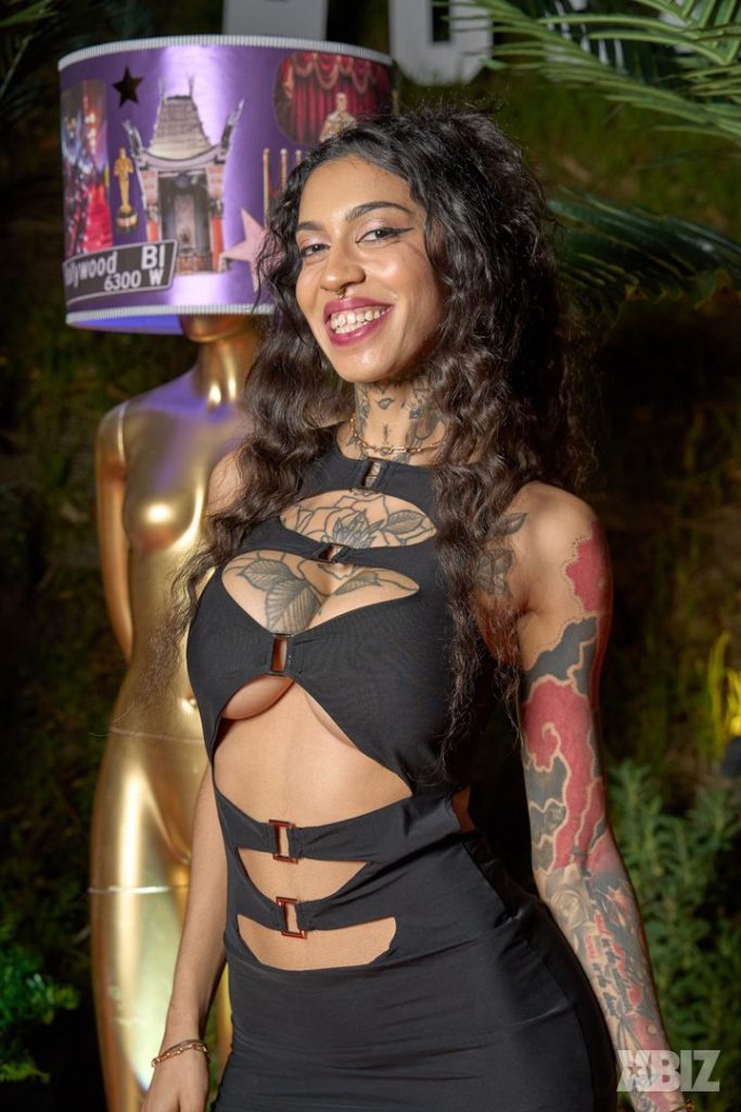 A smiling woman with long curly hair, tattoos on her arms and chest, and wearing a cutout black dress stands outdoors. Behind her is a gold statue and a lamp decorated with Hollywood-themed images.