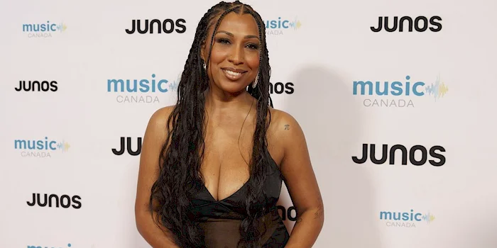 Melanie Fiona in a black dress with long braided hair poses on the red carpet at the JUNOS standing in front of a backdrop with JUNOS and music CANADA logos.
