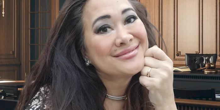 Asian woman with long dark hair smiles, resting her chin on her hand, wearing a patterned top and choker necklace, in a wood-paneled office or courtroom setting.