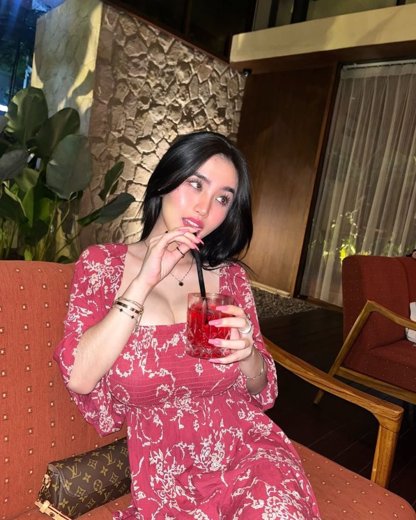 Asian woman in a red floral dress sits on an orange chair indoors, holding a red drink with a black straw and looking to the side. There are plants, stone walls, and large windows in the background.