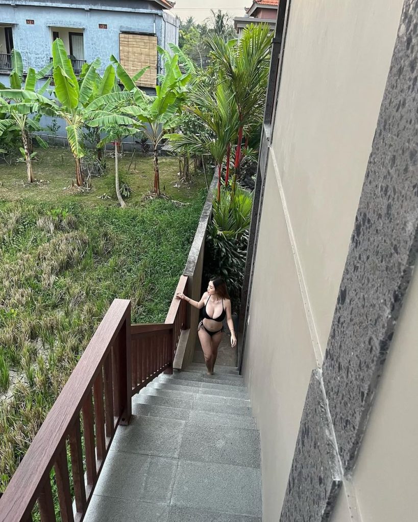 Asian woman in a black bikini walks up outdoor stairs next to a concrete wall, with tropical plants and green grass in the background.