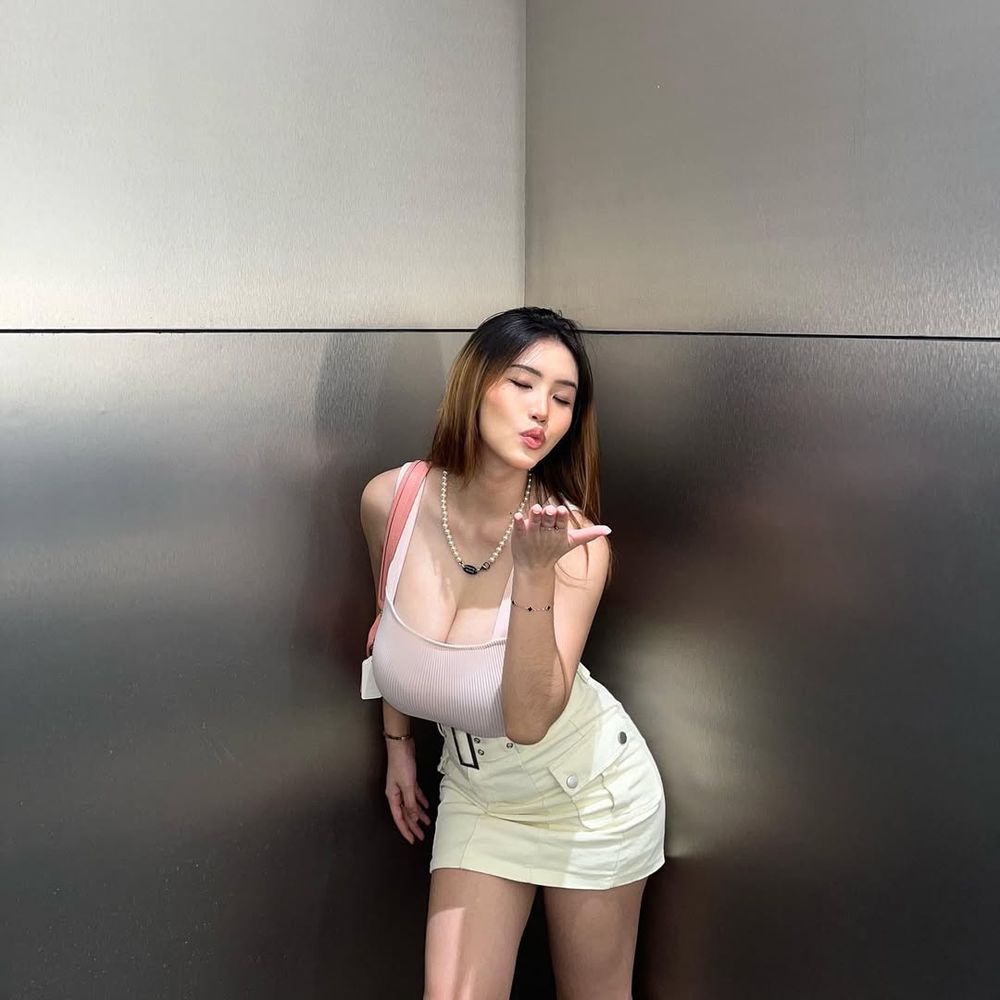 A young Asian woman with long hair wearing a light pink top and cream skirt blows a kiss toward the camera. She stands against a metallic gray background, leaning forward slightly with one hand on her thigh.