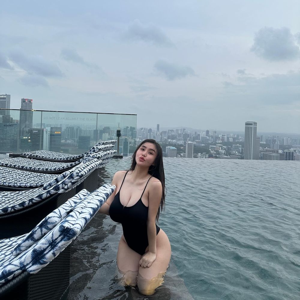 Asian woman in a black swimsuit poses in an infinity pool with a city skyline in the background. She is standing in shallow water near poolside loungers under a cloudy sky.