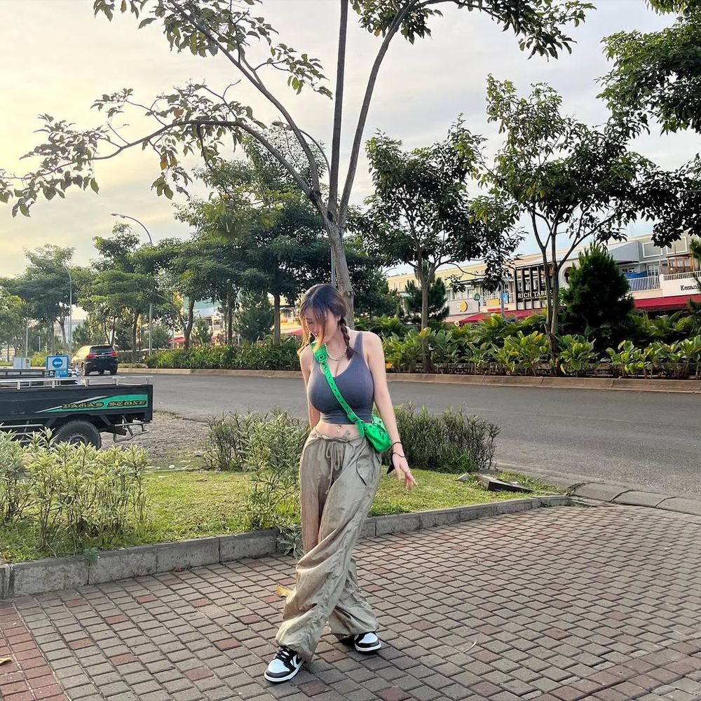 Asian woman with long hair stands on a brick sidewalk near a road, wearing a gray tank top, beige pants, black-and-white sneakers, and a bright green crossbody bag. Trees and bushes line the background under a cloudy sky.