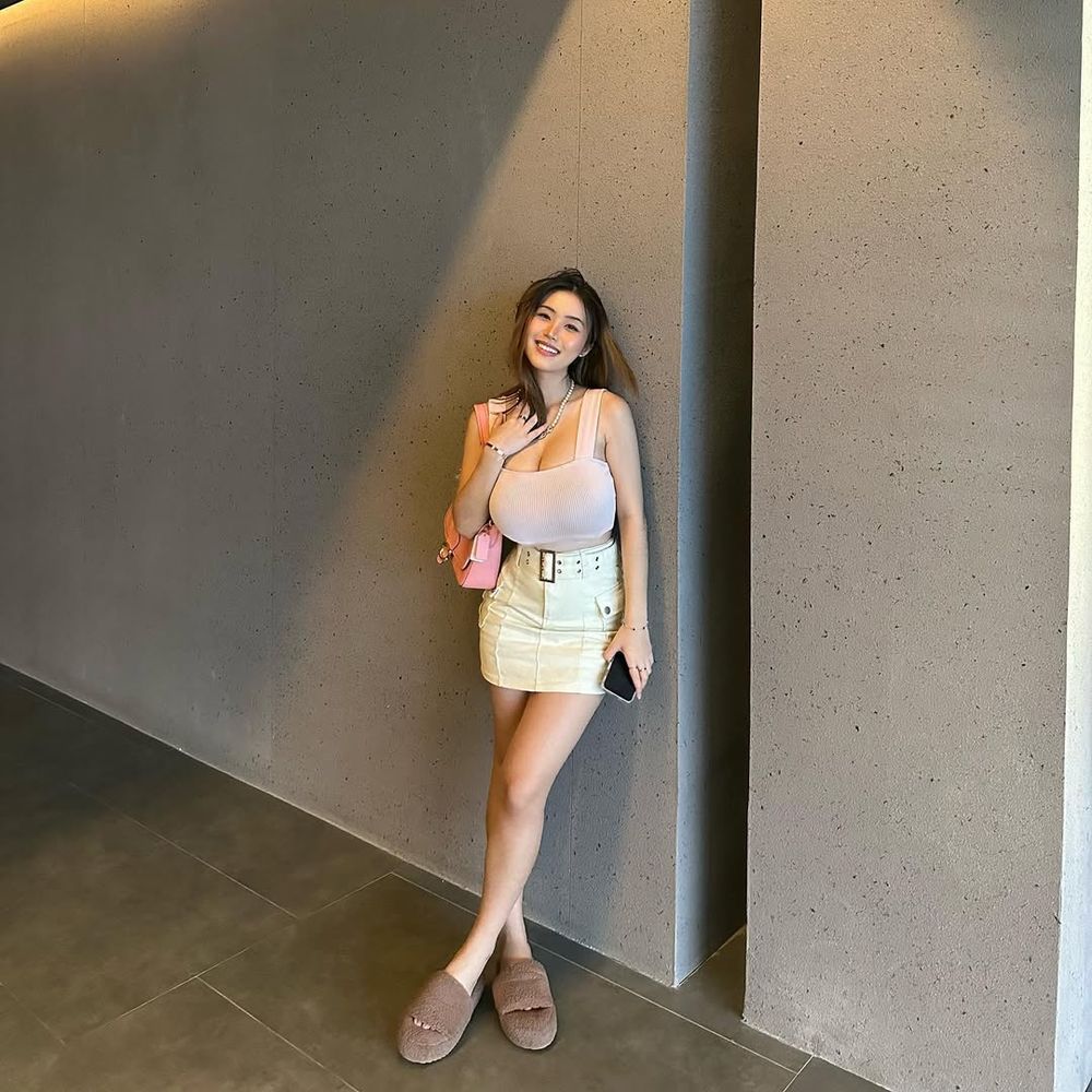 Asian woman stands smiling against a plain textured wall, wearing a light pink sleeveless top, cream mini skirt, brown slippers, and holding a phone. She has a small pink purse slung over her shoulder.